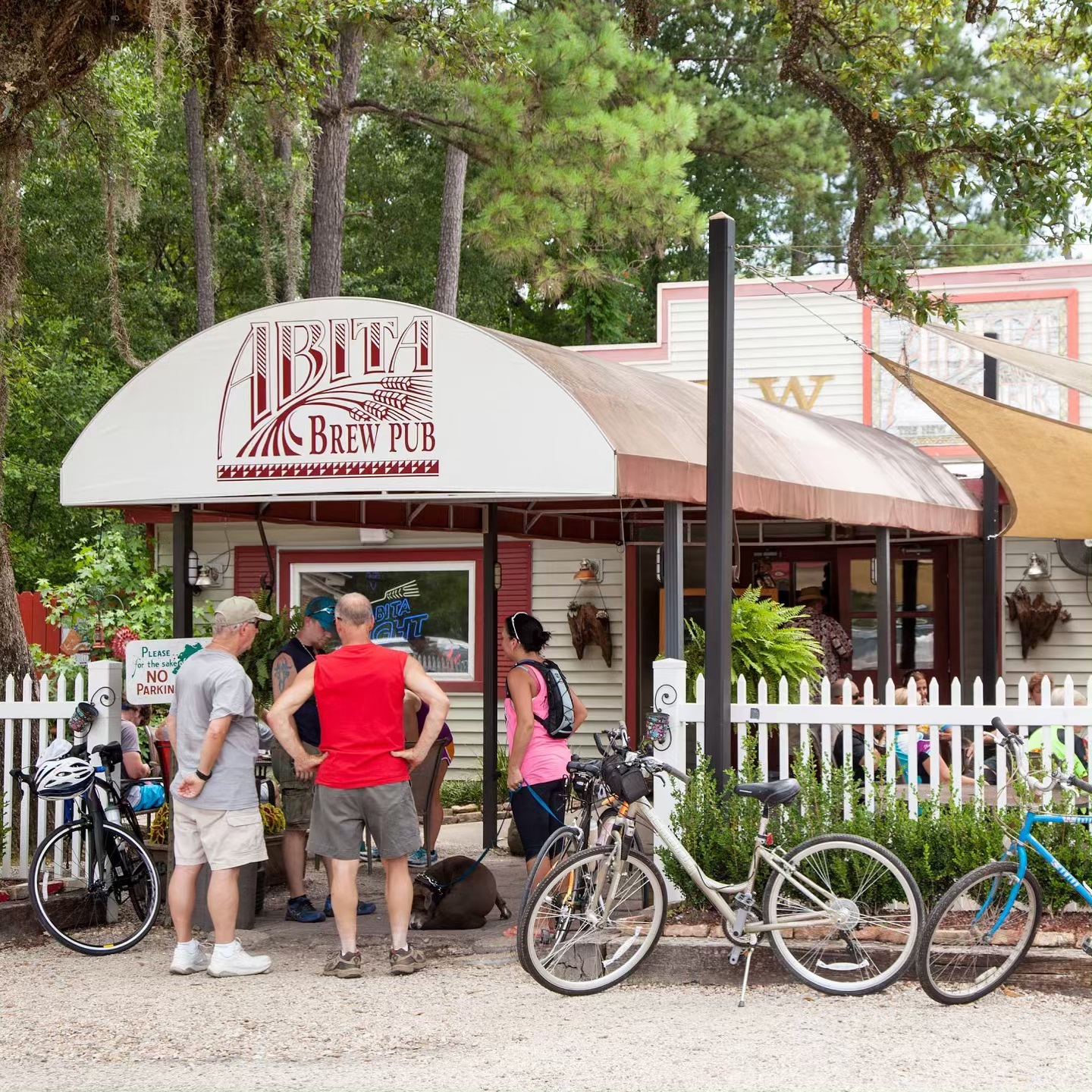 Three people stand underneath an awning with a logo that reads, "Abita Brew Pub." Their bicycles are leaned against a white picket fence to the right of the awning.