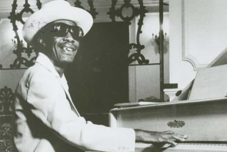 Longhair, in a light-colored suit, straw hat and sunglasses, plays a white piano