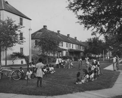 A black and white image of two housing blocks. On the lawn two dozen people lounge.