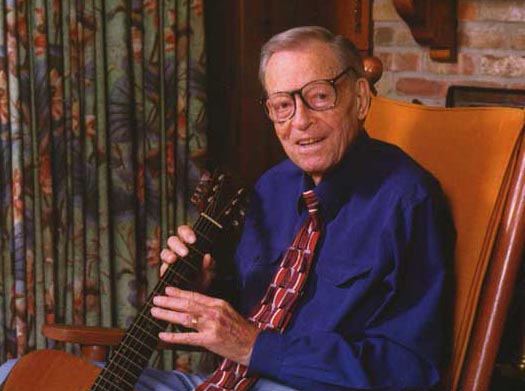 A color photo of Jimmie holding a guitar while sitting in a rocking chair. Curtains and a brick wall are behind him. He wears a navy dress shirt and patterned tie