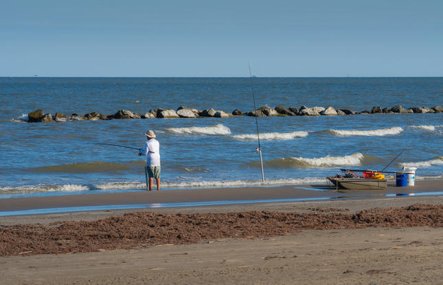 A man stands at the edge of the water holding a fishing rod at Grand Isle State Park, while his boat and other equipment sit just beyond the water.