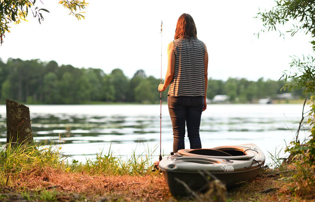 A person in a striped shirt and black pants stands on the shore holding a fishing pole next to a kayak at Jimmie Davis State Park.