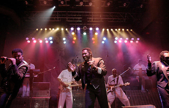 The Commodores perform on stage. Multicolored stage lights illuminate the lead singer.