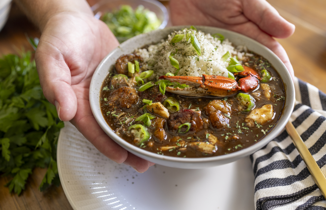 Hands hold a white bowl of gumbo, filled with a brown stew topped with rice, green onions, seafood and other meats.