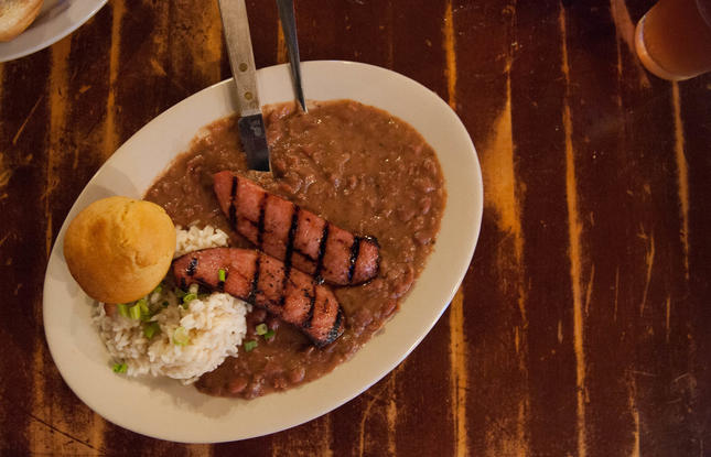 A plate of red beans, topped with sausage scored with grill marks, rice and a piece of cornbread.