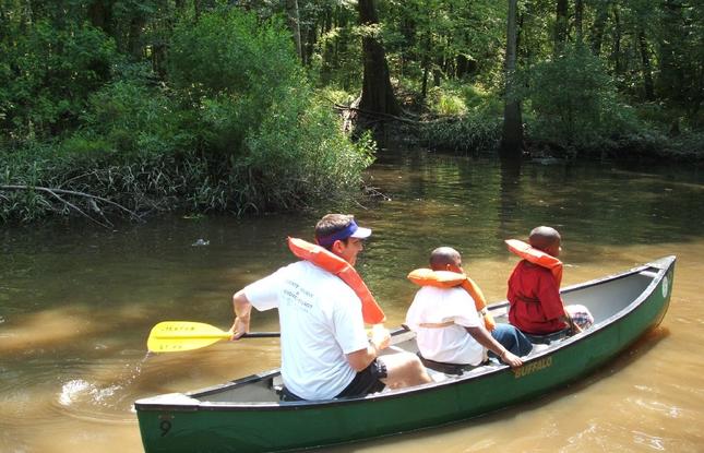 A man and two children wearing lifejackets paddle a canoe through the muddy waters of Tickfaw State Park.