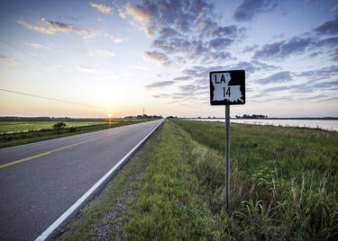 A rural two-lane highway, part of Louisiana's Cajun Corridor Byway, stretches into the distance. In the background the sun peeks over the horizon, brightening a cloudy sky.
