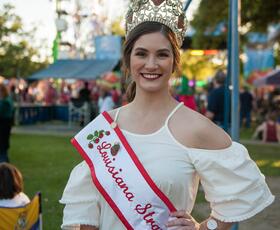 The Louisiana Strawberry Queen poses for a photo in full regalia, with a carnival in the background.