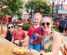 A group of adults and children smile at an outdoor festival table while sharing funnel cake and snacks, with food stands and crowds in the background.
