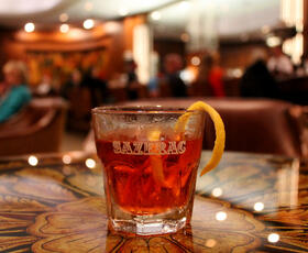 A classic Sazerac cocktail garnished with a lemon twist, served in a glass etched with “Sazerac” at a cozy bar.