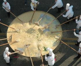Several chefs dressed in white coats and chefs hats stir a massive skillet of eggs at the Giant Omelette Festival.