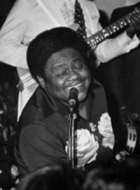 Fats Domino leans away from the piano and into the mic, singin with his eyes closed. Someone behind him plays the guitar.