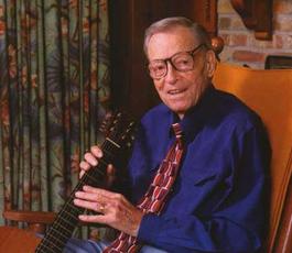 A color photo of Jimmie holding a guitar while sitting in a rocking chair. Curtains and a brick wall are behind him. He wears a navy dress shirt and patterned tie
