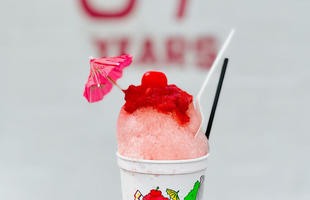 A pink and red snoball fills a cup, and is topped with a small drink umbrella.