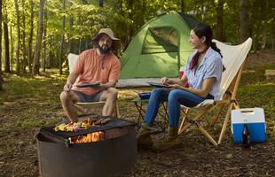 Couple cooking skewers over a campfire beside a green tent in a wooded Louisiana campsite.