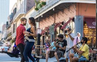 A couple dances in the street while a small brass band plays on Royal Street in New Orleans, with musicians seated and pedestrians watching in the French Quarter.