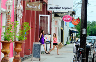 Two women exit a store holding shopping bags on a street lined with colorful shops, including Hemline and American Apparel.